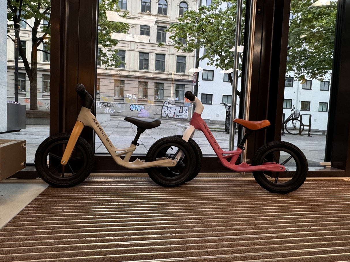 To Mechanic Arts Trillr barnesykler i beige og rosa side om side i butikkvindu – søte balanse sykler for søsken eller venner